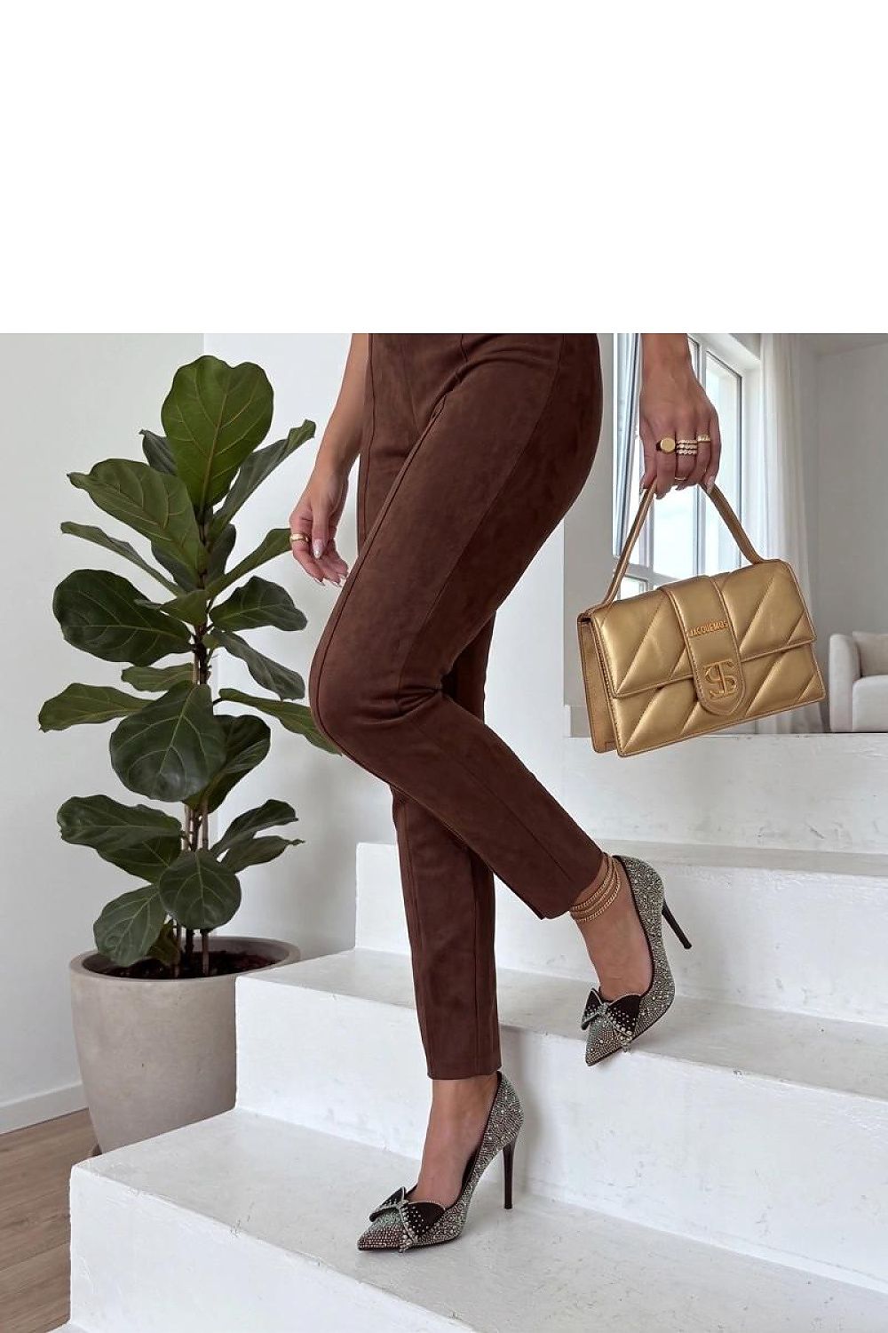 Woman wearing crystal high heels and brown pants, holding a gold handbag, standing on white stairs.